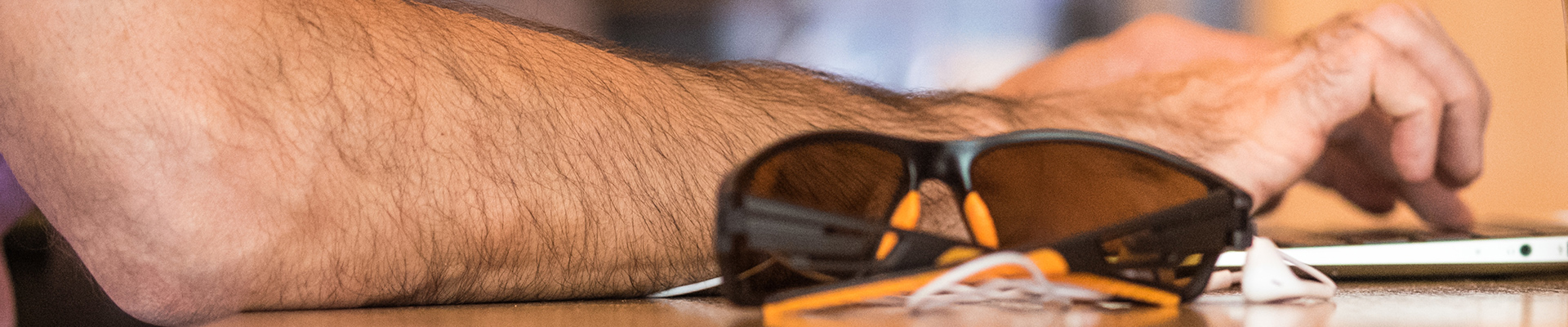 Hand typing on a laptop keyboard with sunglasses and ear buds on the desk