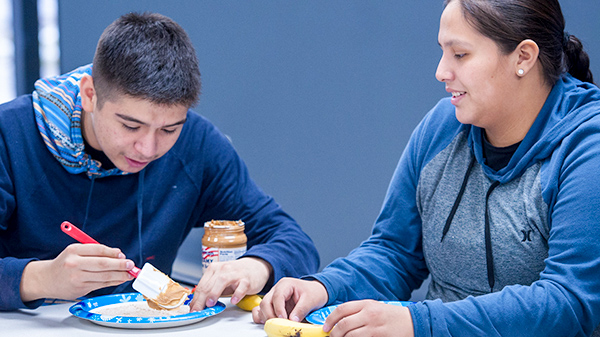 Two young adults sitting at a table, one of them spreads peanut butter on a banana
