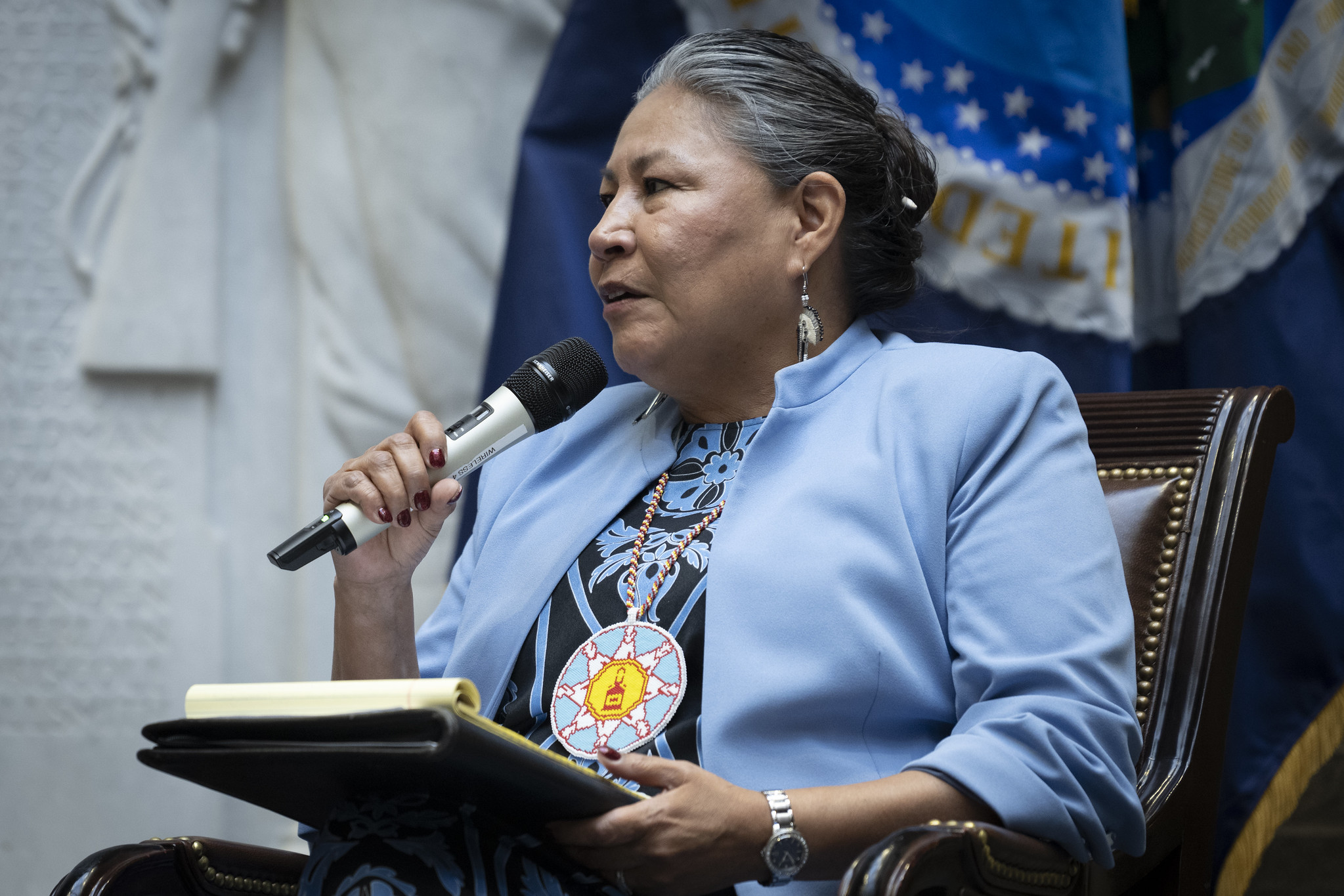 Woman speaking into a microphone while seated at a formal meeting, wearing a blue jacket and beaded medallion.