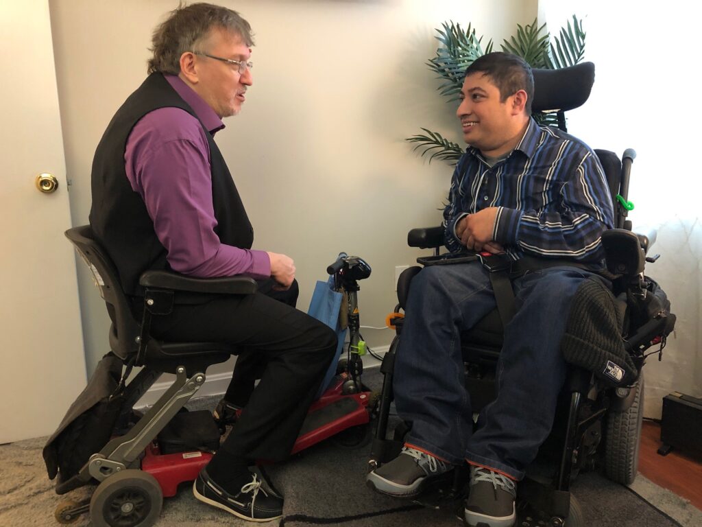 Two people using mobility devices sit facing each other and talking in an indoor setting.