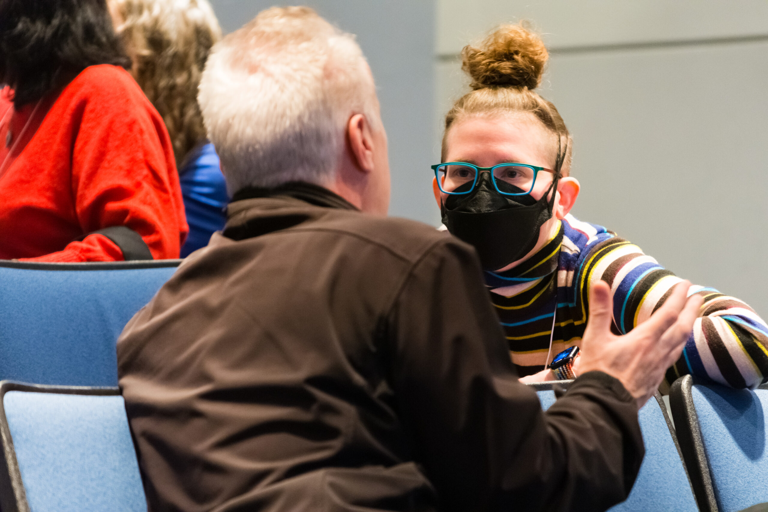 Masked person with glasses speaks with another seated person.