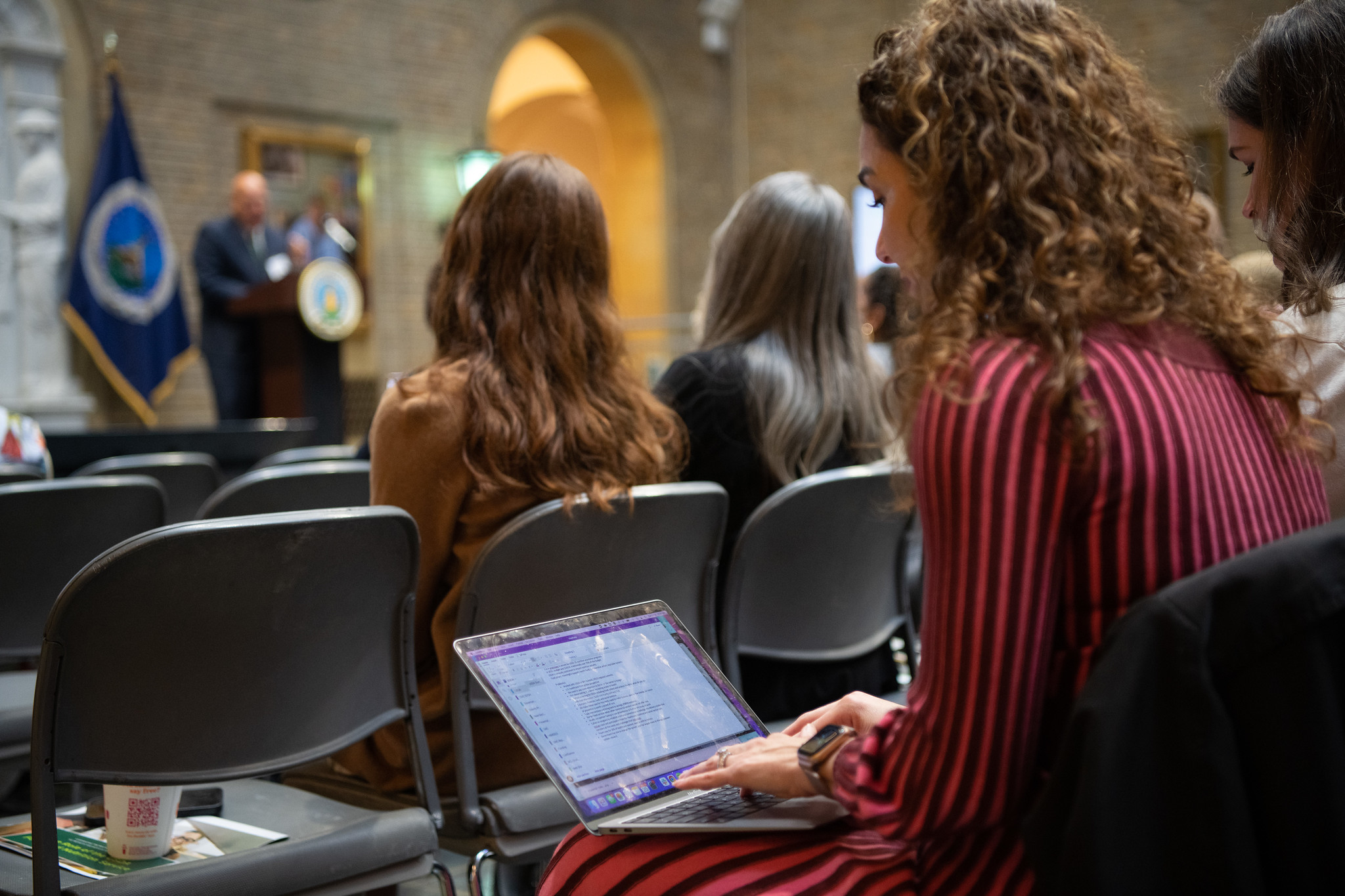 People seated at a public meeting, with one person typing on a laptop while listening to a speaker at a podium.