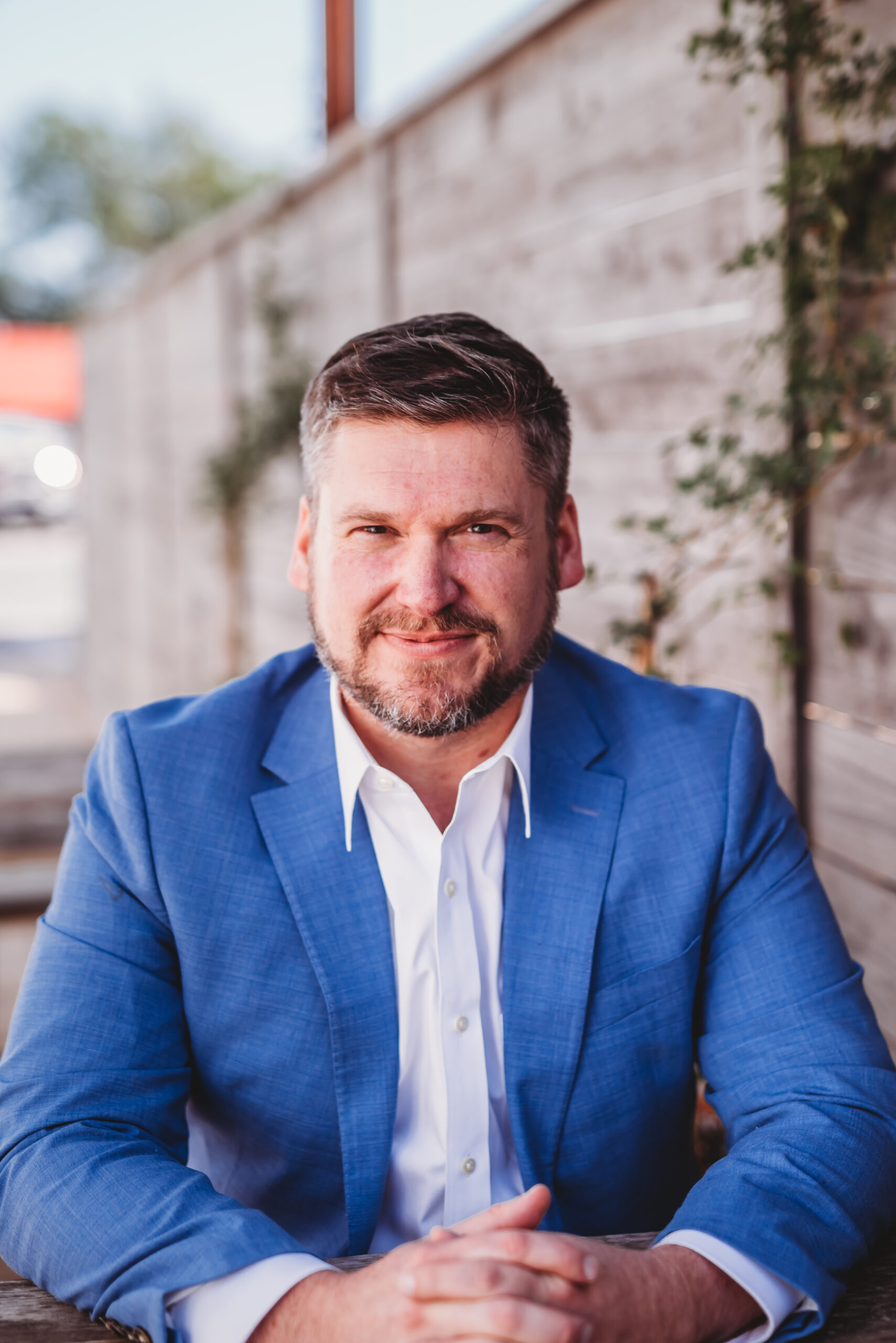 A man in a light blue suit and white shirt sits outdoors, smiling confidently at the camera. A wooden fence and greenery create a warm, natural background