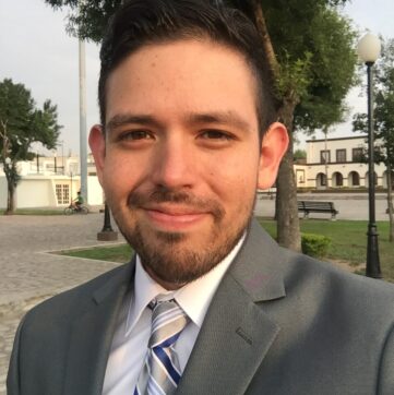 Joey Vega wears a suit and tie with a slight beard and stands outside with trees and a park behind him.