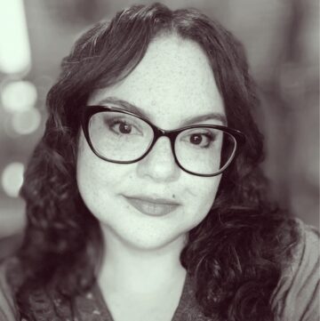 Sandra Breitengross Bitter wears large rimmed, dark, glasses. She has shoulder-length dark hair and dark brown eyes. She is smiling slightly.