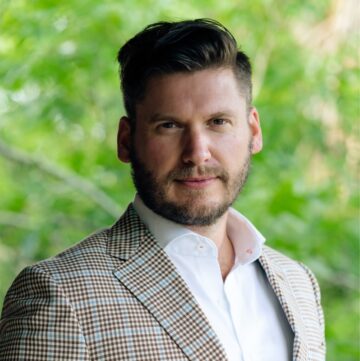Tyler Morris outdoors wearing a plaid blazer and white shirt with a green foliage background.